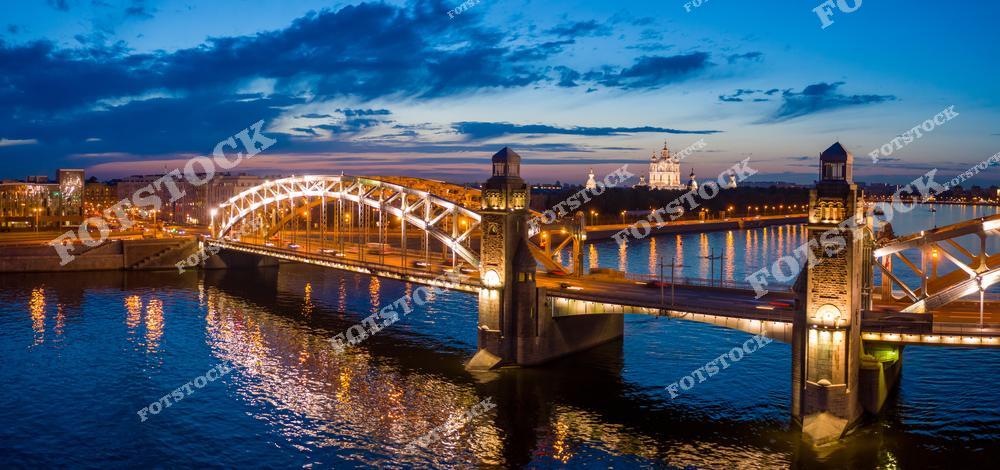 На снимке изображён Большеохтинский мост, великолепно подсвеченный в вечернее время, и отражения его огней в водах Невы. На заднем плане возвышается Смольный собор, добавляя историческую нотку к современному мосту. Красочное небо на закате создаёт уютную и живописную атмосферу. Это одна из самых известных панорам Петербурга, соединяющая архитектурную классику и современные инженерные решения.